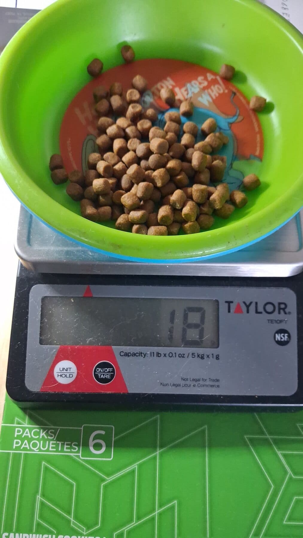 Un tazón verde con croquetas encima de una báscula de cocina que marca 18 gramos.